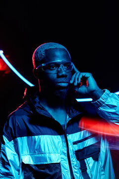 Vertical medium portrait of African American guy with blond hair wearing stylish outfit and futuristic AR eyewear standing in dark studio room in neon lights touching button on eyeglasses