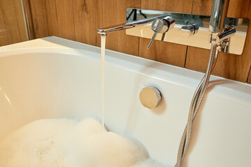 Modern chrome bathtub faucet pouring water into tub. Concept of home wellness, self care and relaxation in bathroom