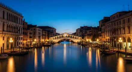 Naklejka premium Photo of the rialto bridge spans the grand canal in venice, italy, illuminated by warm lights under a clear blue evening sky