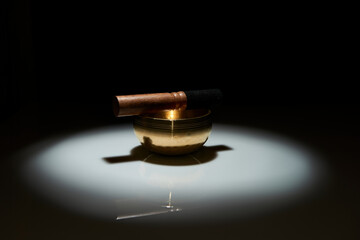 A Tibetan singing bowl and mallet illuminated by a dramatic spotlight against a dark background,...