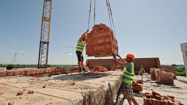 Construction worker guiding brick pallet lifted by crane. Worker in safety gear directs a large pallet of hollow red bricks being lifted by crane at a building site on a sunny day.