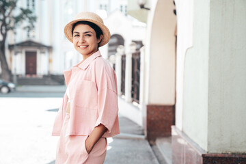 Obraz premium Young beautiful smiling hipster woman in trendy summer pink costume clothes. Carefree female posing in the street at sunny day. Positive model outdoors at sunset. Cheerful and happy in hat