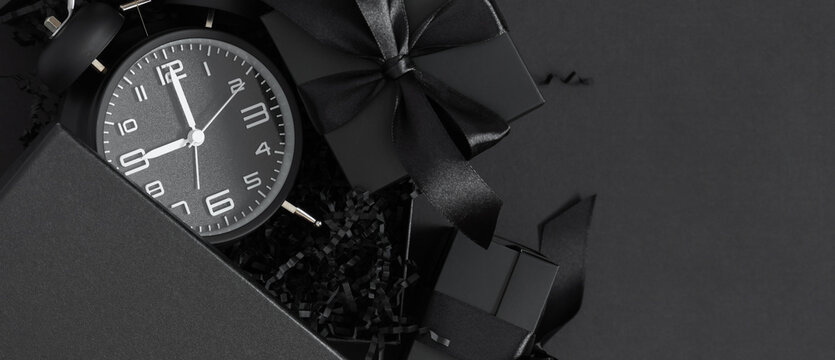 Black gift box and black alarm clock on dark background is perfect for Black Friday shopping banner design.