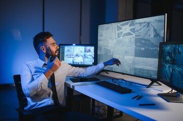 Male Security Guard Talking On Walkie talkie While Monitoring Multiple CCTV Footage On Computer