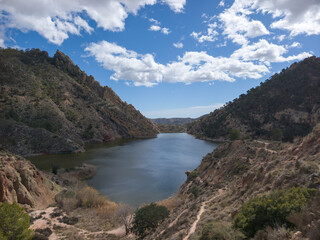 Obraz premium Serene reservoir nestled between rocky mountains under a bright blue sky with fluffy clouds, perfect for outdoor recreation