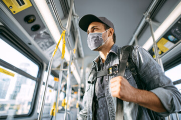 Man wearing face mask, casual clothing, and cap standing inside city bus, holding backpack strap, urban public transportation