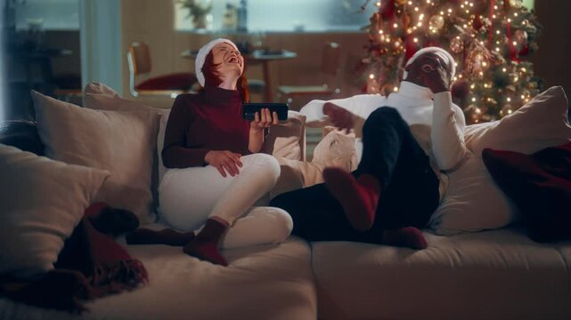 Cozy evening at home in Christmas holidays, biracial lovers relaxing on sofa. Happy young Caucasian woman and her African American boyfriend sitting on soft couch and talking about funny memes in web