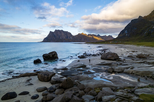 Aerial view of a solitary figure wanders along a rocky beach under the vast, cloud-strewn sky near jagged peaks, Loften Islands, Nordland, Norway.