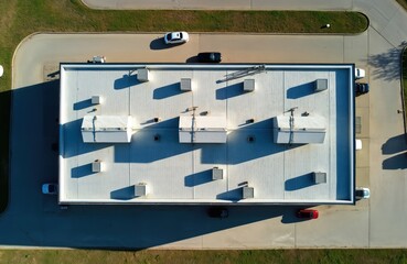 Top aerial view of modern industrial building flat shingle roof. Features include various ventilation systems, pipes, AC units casting long shadows on clean surface. Surrounding asphalt, green grass