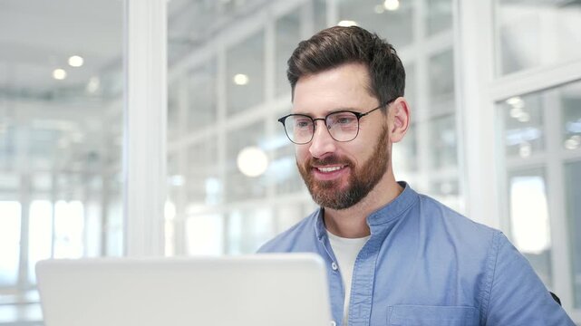 A smiling businessman working on a laptop sitting at workplace in business office. Handsome bearded male worker typing on a computer, texting a client, chatting online or busy with a project. Close up - Powered by Adobe