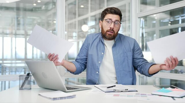 Confused businessman having difficulty with paperwork sitting at workplace in business office. Puzzled disappointed entrepreneur looks at documents, cannot understand the problem and looking at camera