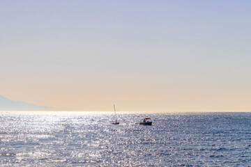 Minimalistic seascape with a lone sailing boat gliding across the ocean, illuminated by the shimmering sunlight of a golden sunset. The soft pastel tones of the sky and gentle reflections on the water
