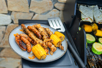 Grilled sausages, skewers, and corn on a plate near a gas barbecue with vegetables and flatbreads cooking. Outdoor summer cooking scene with delicious grilled food.