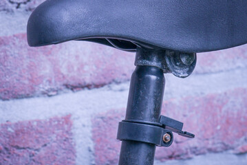Close-up of an old, rusty adjustable black saddle mounted on a sturdy bike seat post against a brick wall