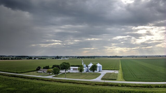 Aerial view of sun rays breaking through the clouds, illuminating the silos and farm amidst the vast green fields, Monroeville, Indiana, United States.