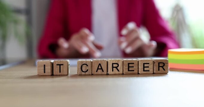 Wooden blocks with IT Career on desk with woman in pink blazer typing on keyboard. Pursuing career in information technology in office setting
