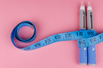 Overhead shot of semaglutide pens wrapped around measuring tape on pink table, symbolizing weight...