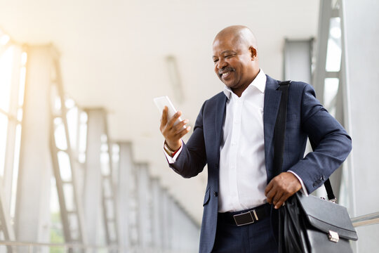 Happy black businessman in suit smiling at smartphone inside airport terminal. Corporate travel, modern communication, mobility and professional global lifestyle