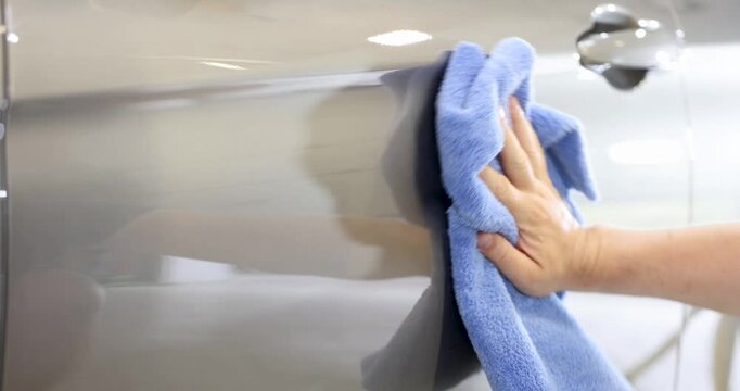Hand vigorously rubs vehicle surface with blue microfiber cloth during cleaning. Pressure intensifies effort creating visible shine on car paint
