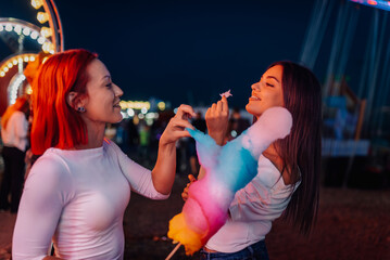 Two young women sharing cotton candy at amusement park by night