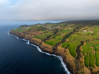 Aerial view of the rugged coastline where the dark blue sea crashes against the layered cliffs beneath vibrant green fields, Ginetes, AÃ§ores, Portugal.
