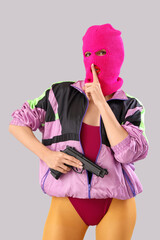 Beautiful young stylish woman in balaclava with gun showing silent gesture on grey background