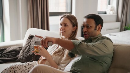 A pregnant woman is holding a cup and talking to a man who is clicking on a remote control while they are sitting on the sofa - Powered by Adobe