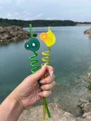 A person holds two bright, twisted straws designed to look like fruit, with a picturesque lake and...