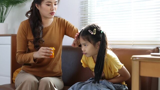Nurturing Asian mother providing comfort and administering medication to her ill daughter experiencing stomach pain while sitting close together on living room sofa.