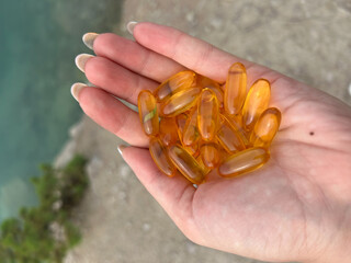 A hand displays a variety of orange capsules against a natural backdrop of water and rocks. This...