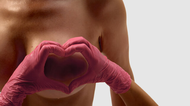 Pink gloves forming heart shape gesture on female chest closeup. Concept of breast health awareness, plastic surgery, medical examination, femininity, wellness, and body care.