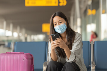 A woman wearing a face mask sits in an airport waiting area, looking at her smartphone, with a pink suitcase beside her on the empty row of blue chairs