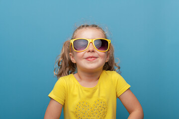 A cheerful baby girl with curly hair wearing yellow sunglasses and a yellow t-shirt poses confidently against a bright blue background in a playful studio portrait.