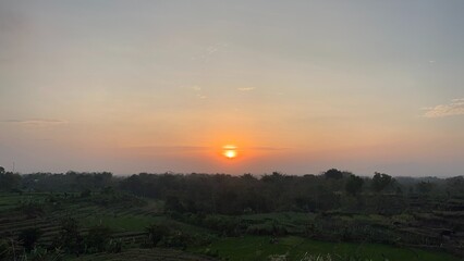 Peaceful sunrise above green farmland with soft orange glow, rural countryside landscape with silhouetted trees and hills.