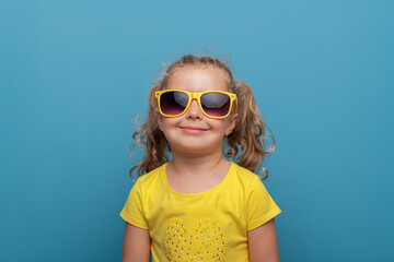 A cheerful baby girl with curly hair wearing yellow sunglasses and a yellow t-shirt poses confidently against a bright blue background in a playful studio portrait.