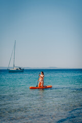 Naklejka premium Woman kneeling on sup board paddling in the sea with sailboat in background