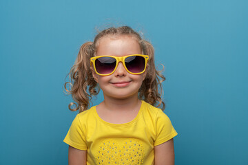 A cheerful baby girl with curly hair wearing yellow sunglasses and a yellow t-shirt poses confidently against a bright blue background in a playful studio portrait.