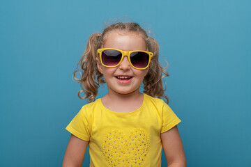 A cheerful baby girl with curly hair wearing yellow sunglasses and a yellow t-shirt poses confidently against a bright blue background in a playful studio portrait.