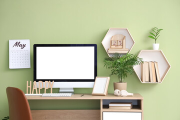 Comfortable workplace with modern PC monitor, headphones, shelves and calendars hanging on color wall