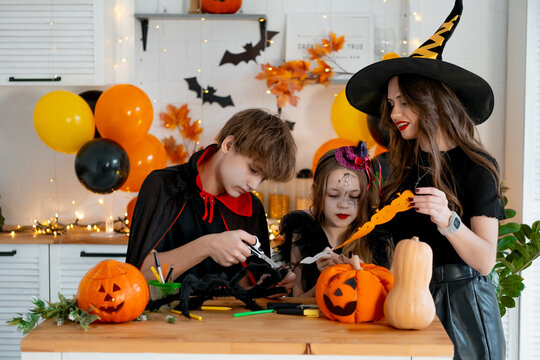 Happy family preparing for Halloween. Mother and children in carnival costumes at home in the kitchen have fun, scaring each other and making or carving pumpkins. - Powered by Adobe