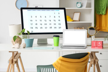 Modern computer monitor with calendar, laptop, headphones and cup of coffee on table in light room