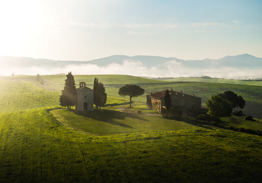 Aerial view of the iconic Cappella della Madonna di Vitaleta standing proudly amidst rolling green hills under a pastel sky, San Quirico d'Orcia, Tuscany, Italy.