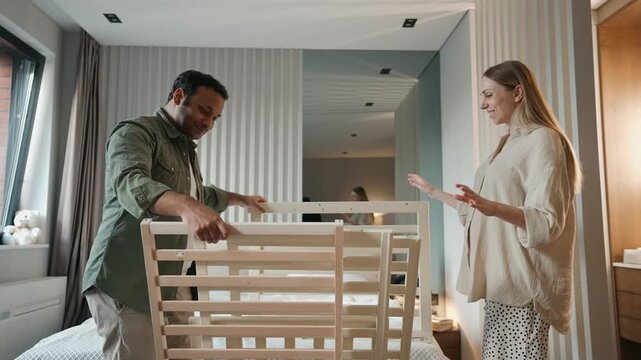 A pregnant woman approaches a man who is holding parts of a crib while they talk