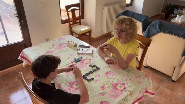 Mother and her 12-year-old son sitting at a floral-covered table playing dominoes indoors, sharing a joyful moment together as the mother claps in celebration during their game