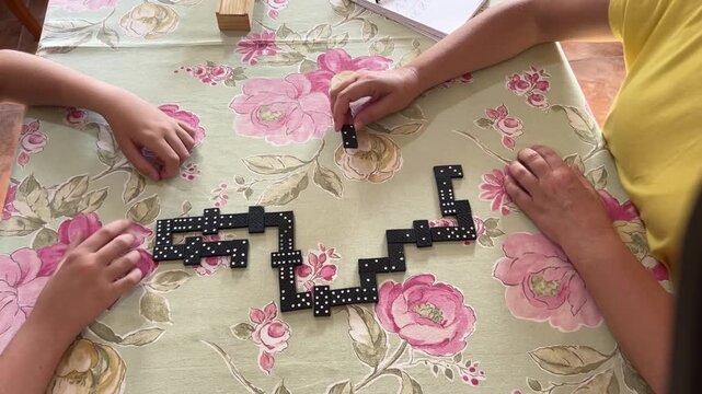 Mother and son playing a strategy-filled game of dominoes on a floral tablecloth, placing pieces with focus and engagement during a relaxed family moment at home