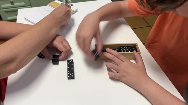 Mother and her 12-year-old son organizing and storing domino pieces in a wooden box on a white table after playing a game, showcasing cooperation and shared responsibility at home