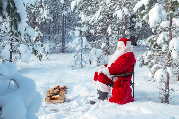 Authentic Santa Claus with a bag of gifts sits near a fire in a snowy forest.