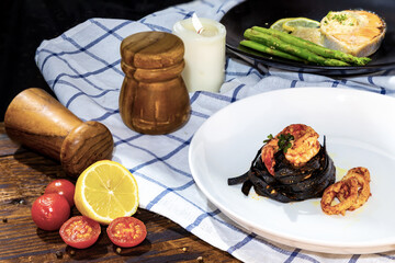 Black fettuccine spaghetti pasta with rock shrimp and squid topping with green parsley; Salmon steak with green asparagus on woodden table, napkin.