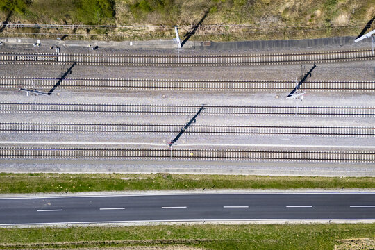 Aerial view of parallel train tracks glinting under the sun, contrasted by the dark asphalt road and bordered by green grassy verges, Deutschlandsberg, Steiermark, Austria.