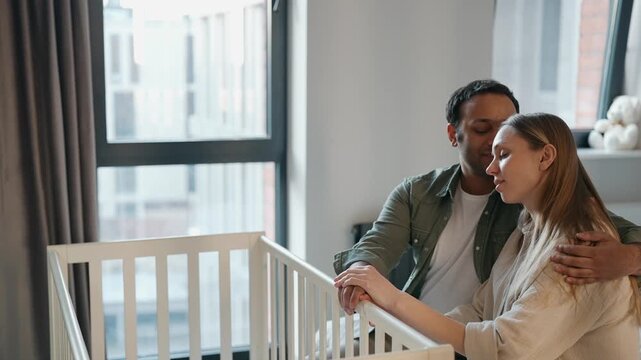 A man hugs and kisses a pregnant woman on the head while they sit on the bed and put their hands on the crib